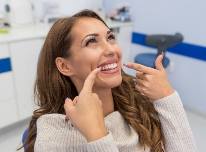 Woman in white sweater pointing to her smile while sitting in dental chair