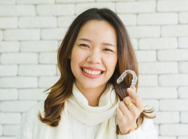 Smiling woman in white sweater holding Invisalign clear aligner